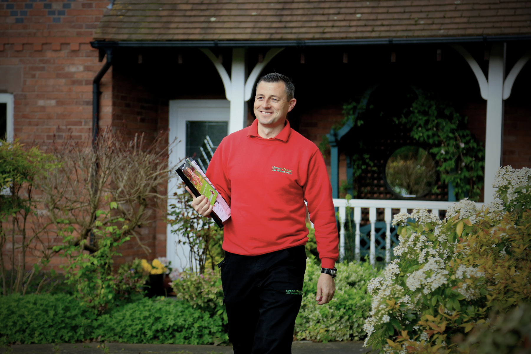 GreenThumb lawn operative in a red uniform walking through a garden, holding treatment products.