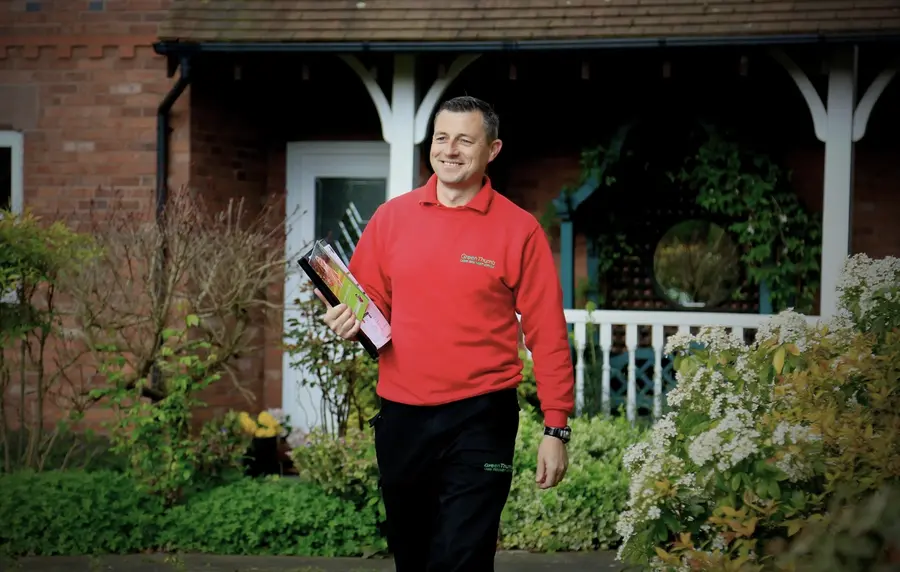 GreenThumb lawn operative in a red uniform walking through a garden, holding treatment products.