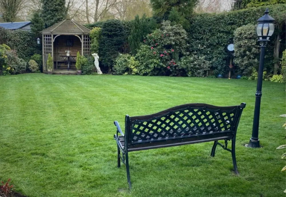 Neatly mown green lawn with a black garden bench and wooden gazebo in autumn/winter.