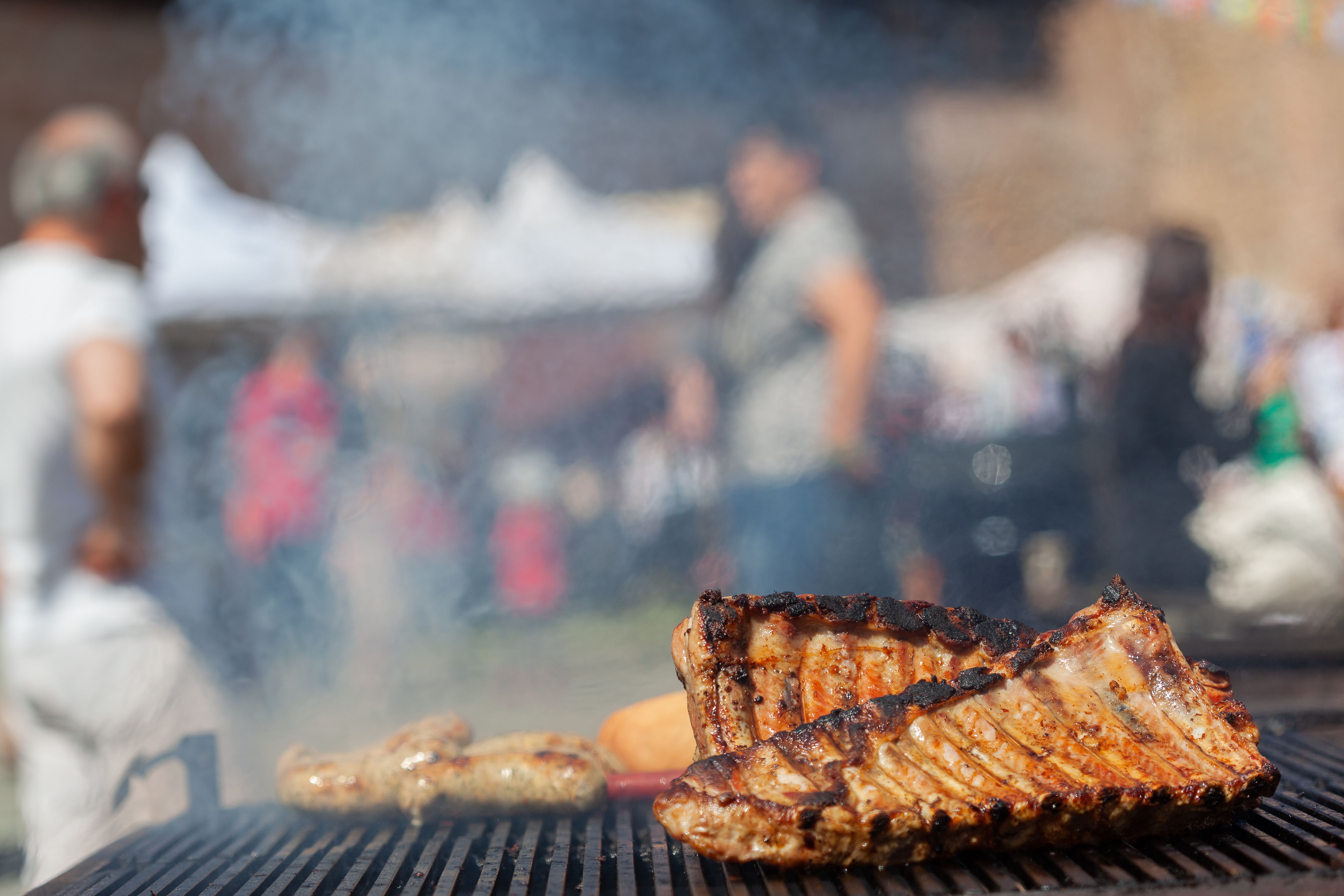 Image Ribs and sausage grilling over an open flame, symbolizing the PSL Bacon and BBQ Fest with blurred festival crowd in the background.