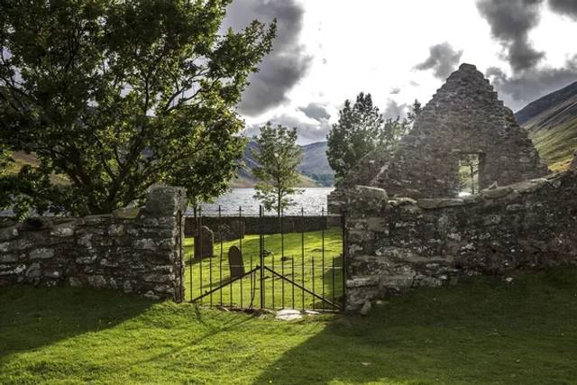 Stone ruins with black metal gate opening to green field, with mountains and a lake visible beyond under a cloudy sky