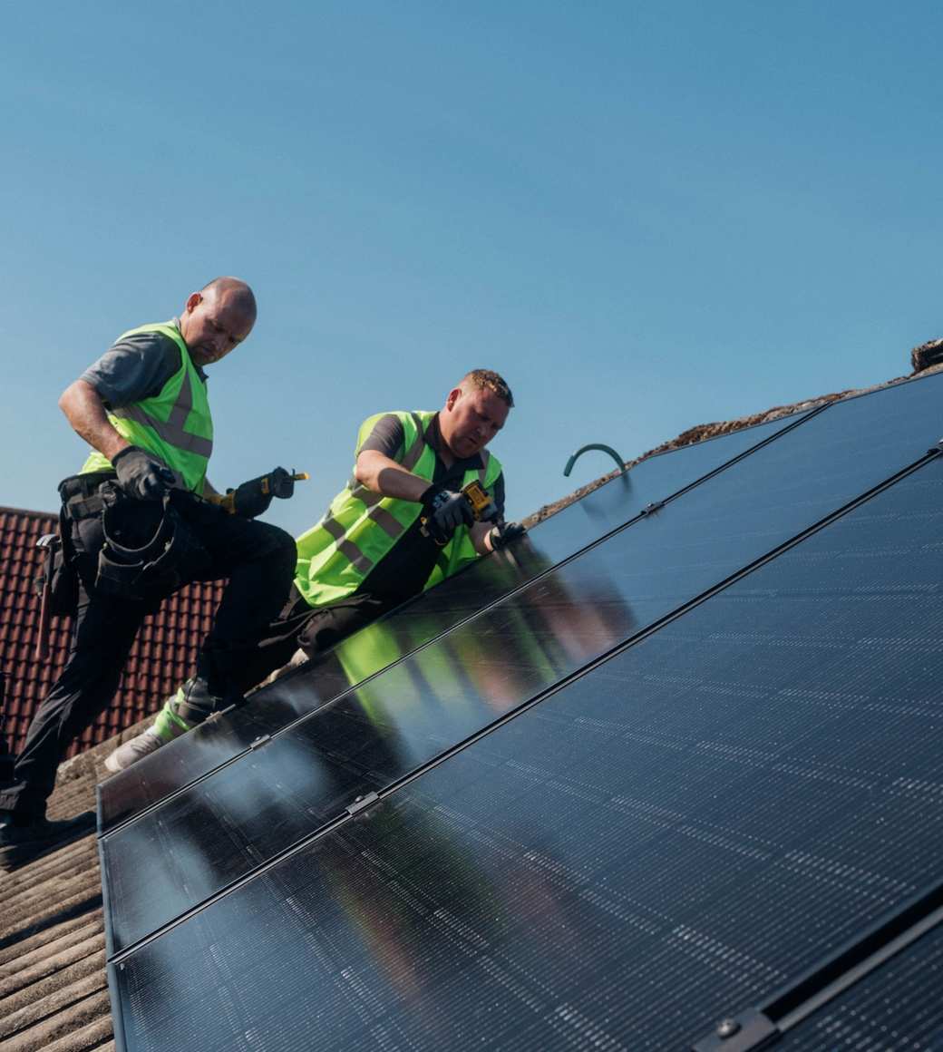 Two workers in high-visibility vests installing solar panels on a residential rooftop under clear blue sky