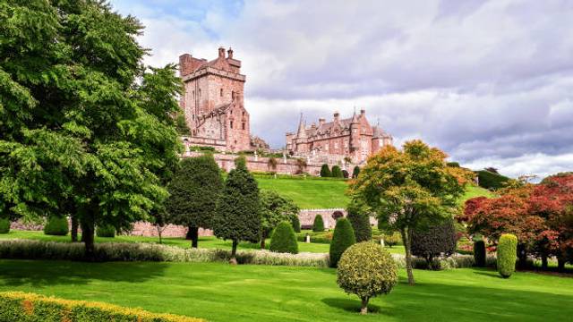 Grand Scottish castle with red stone towers on a hillside, surrounded by manicured gardens with topiary and autumn trees
