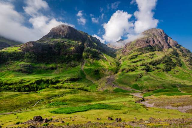 Two rugged mountain peaks with lush green slopes rise against a blue sky, with a small stream running through the valley below