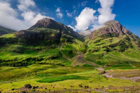 Two rugged mountain peaks with lush green slopes rise against a blue sky, with a small stream running through the valley below