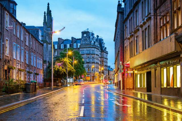 Evening street scene in a Scottish city centre with wet cobblestone road, historic architecture, illuminated storefronts, and a church spire
