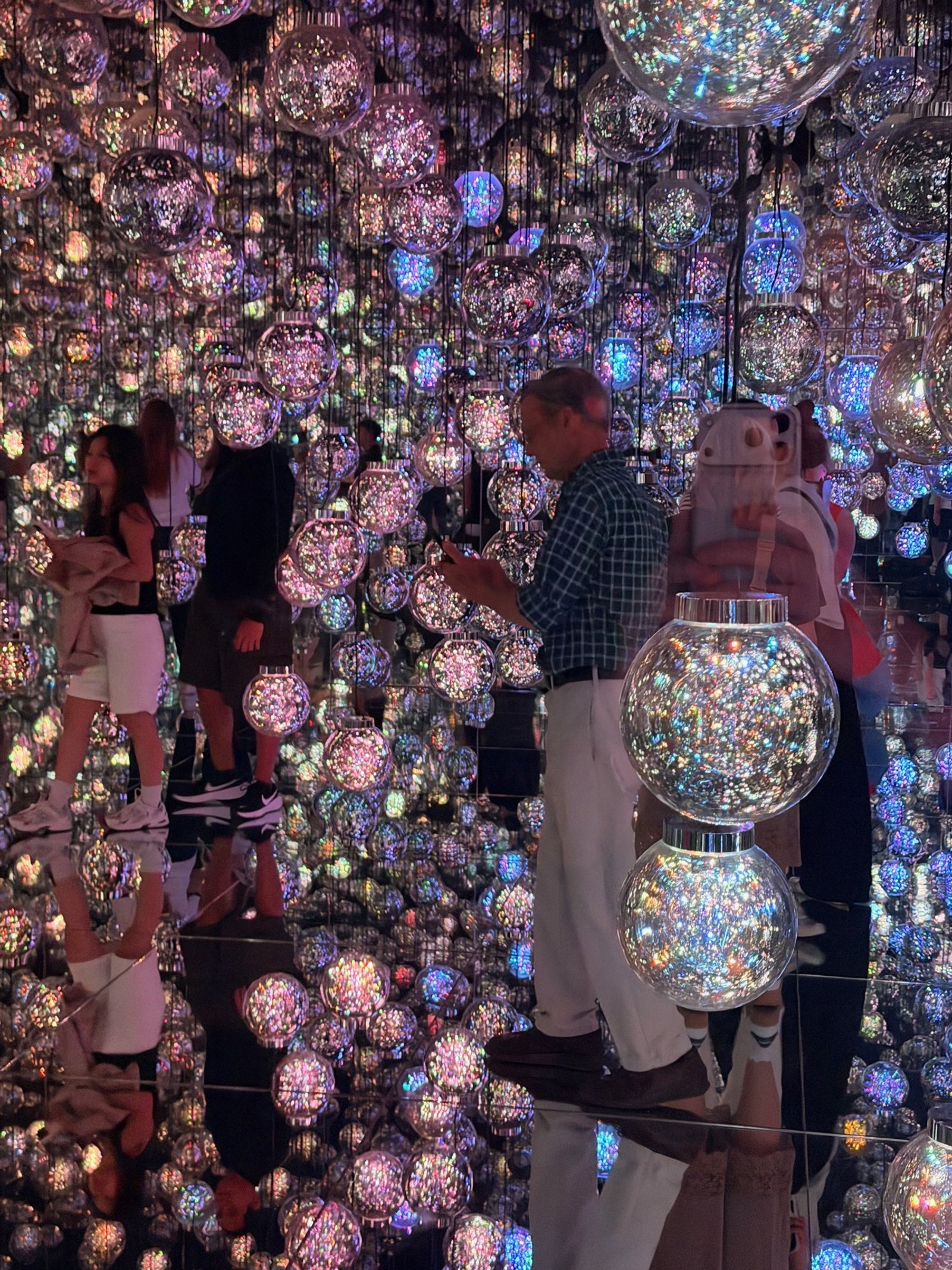 Visitors and hanging illuminated spheres repeated in mirrors inside an immersive art installation.