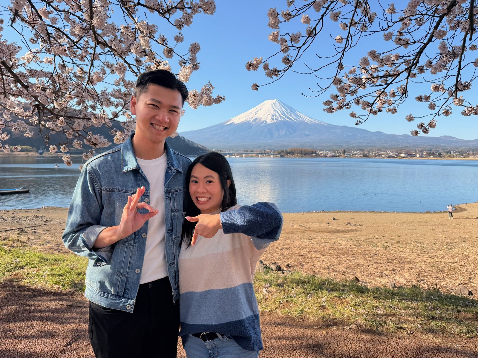 Two smiling travelers in a playful pose beneath blossoms, with a lake and snow-capped Mount Fuji behind them.