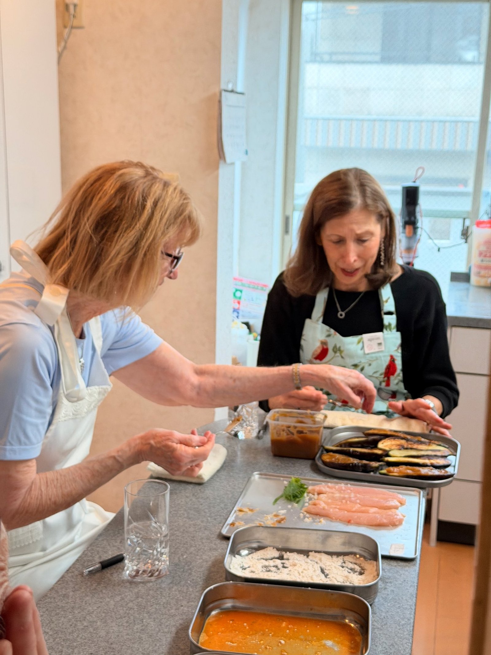 Two participants in aprons preparing food at a kitchen counter with trays of ingredients.