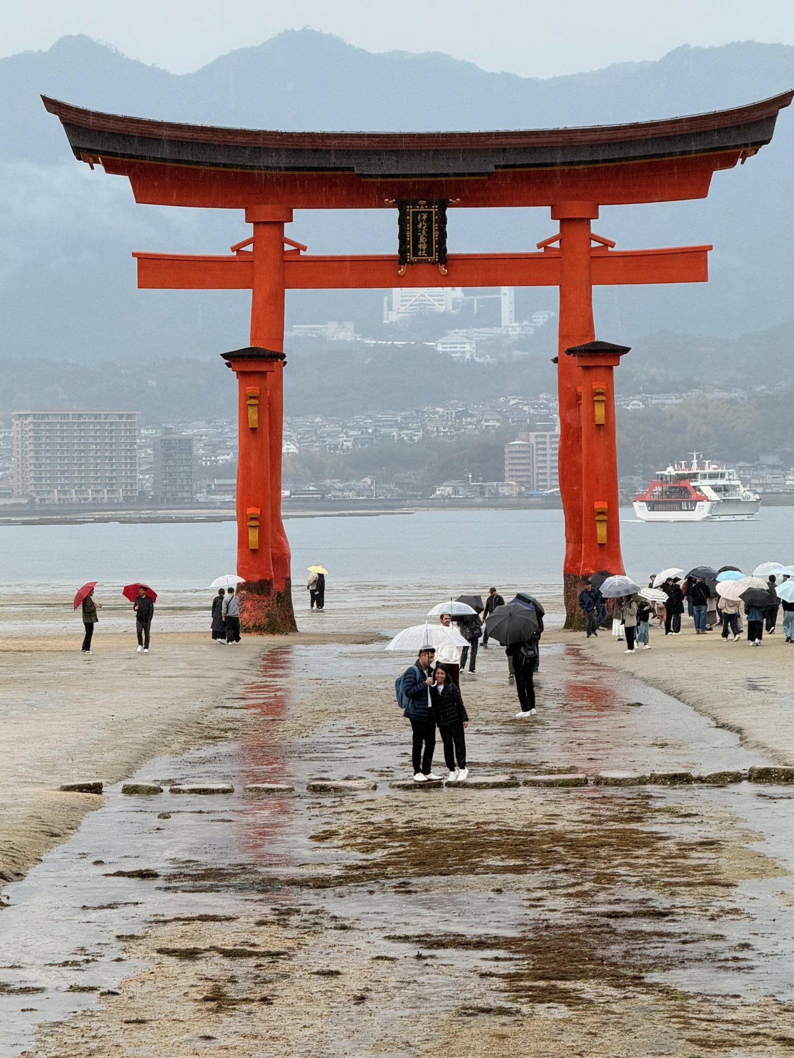 Two travelers beneath an umbrella in front of Miyajima’s enormous red torii, with visitors and umbrellas on wet sand.