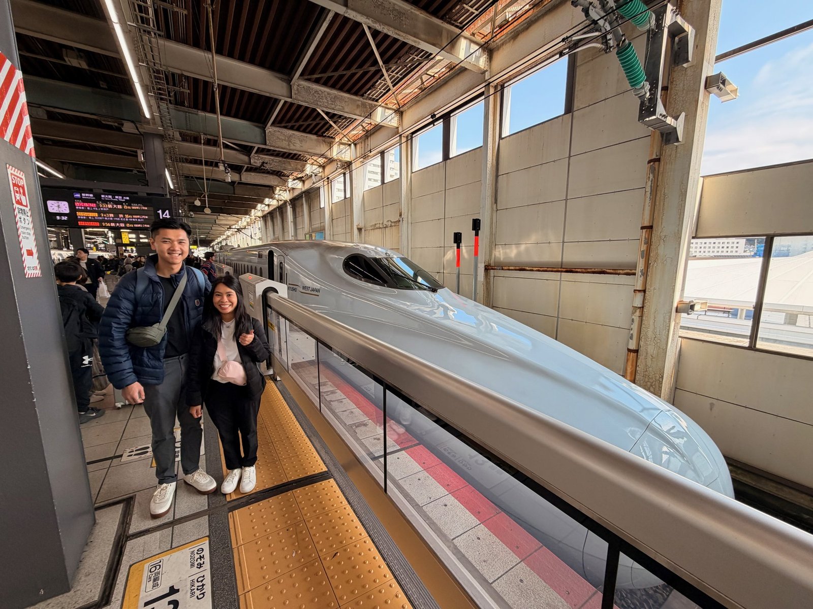 Two smiling travelers on a station platform beside a silver Shinkansen.