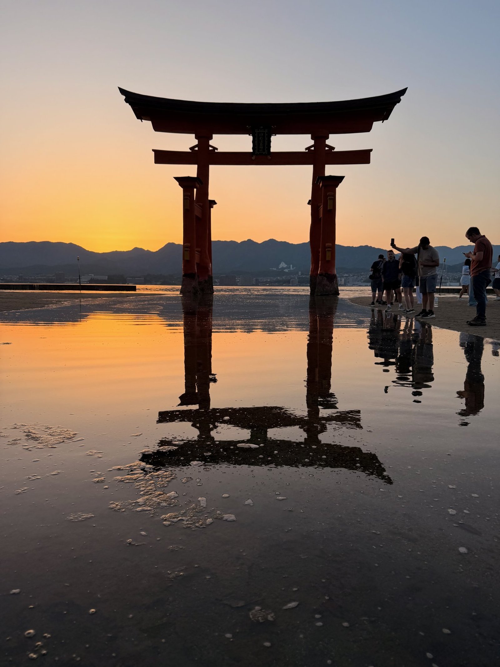 Miyajima’s great torii reflected in shallow water at sunset, with visitors beside it.