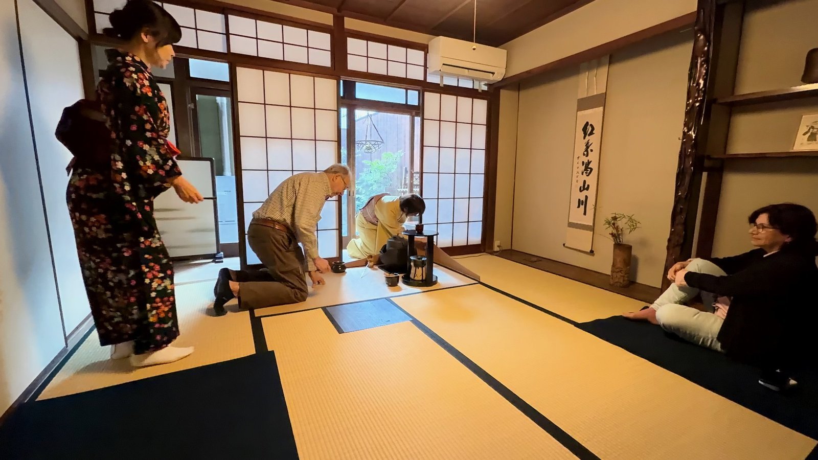 A participant kneeling near tea utensils while a host demonstrates in a tatami room.