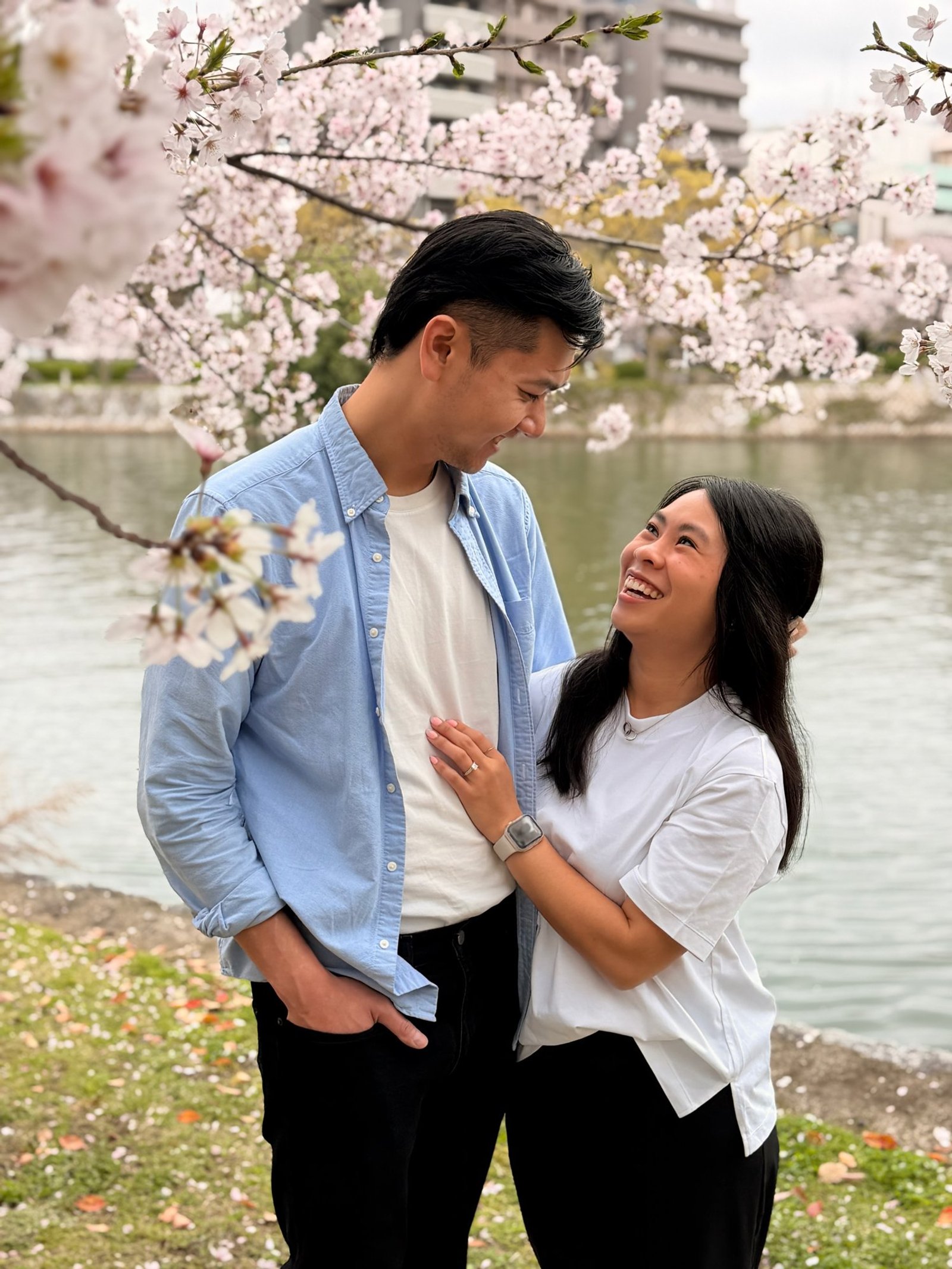Two travelers looking at each other beneath pale cherry blossoms beside a river.
