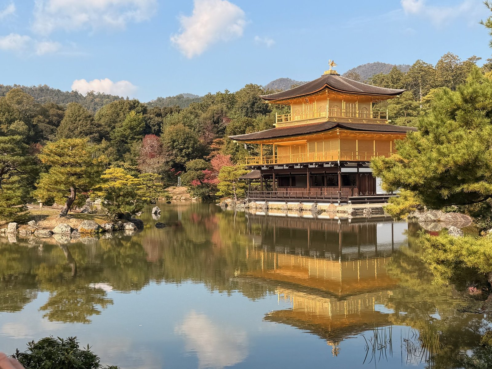Kinkaku-ji reflected in still water, with trees and a blue sky around it.