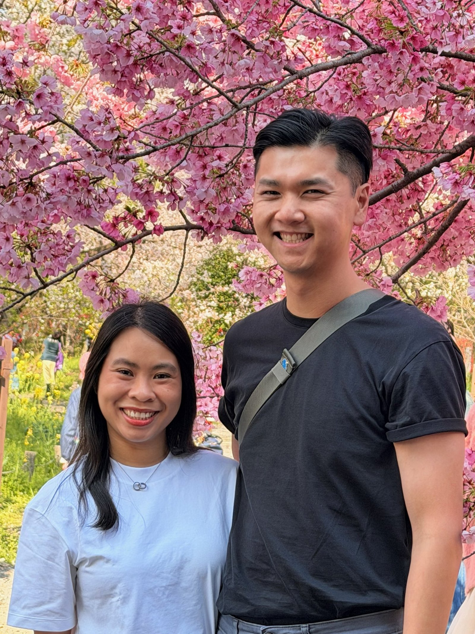 Two smiling travelers standing beneath dense pink cherry blossoms.