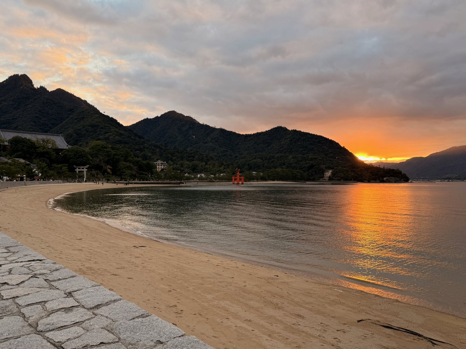 A curving beach and small red torii beneath dark mountain slopes, with orange sunset reflected across the water.