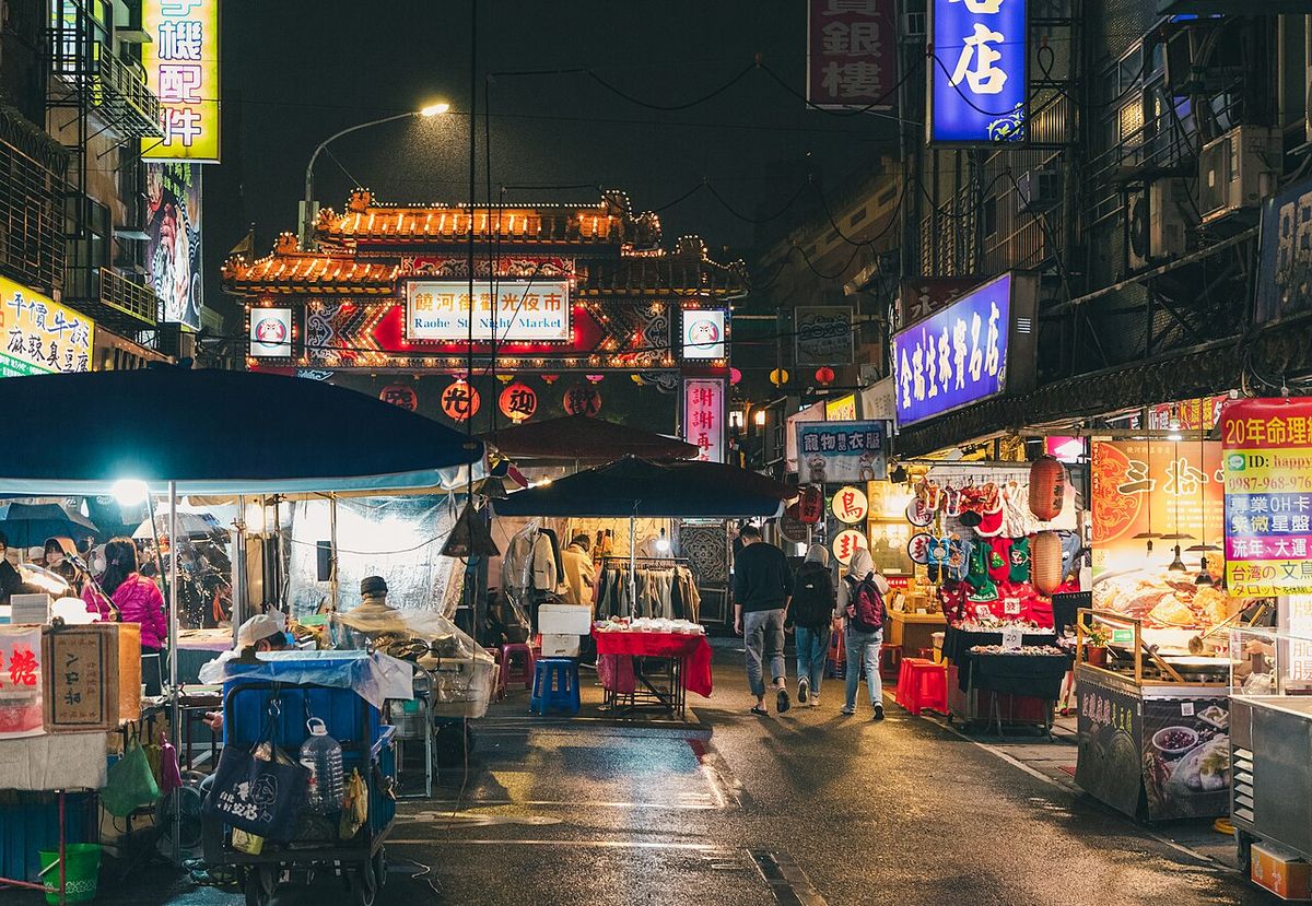 Raohe Night Market street at night with food stalls, lanterns and the lit gate of Ciyou Temple