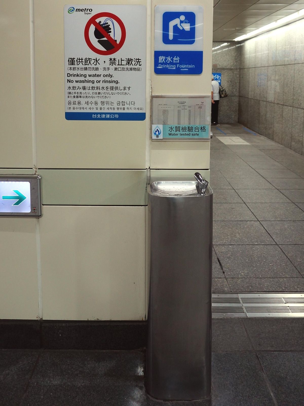 A stainless steel public drinking fountain in Taipei's Ximen MRT station, with a water quality sign above it