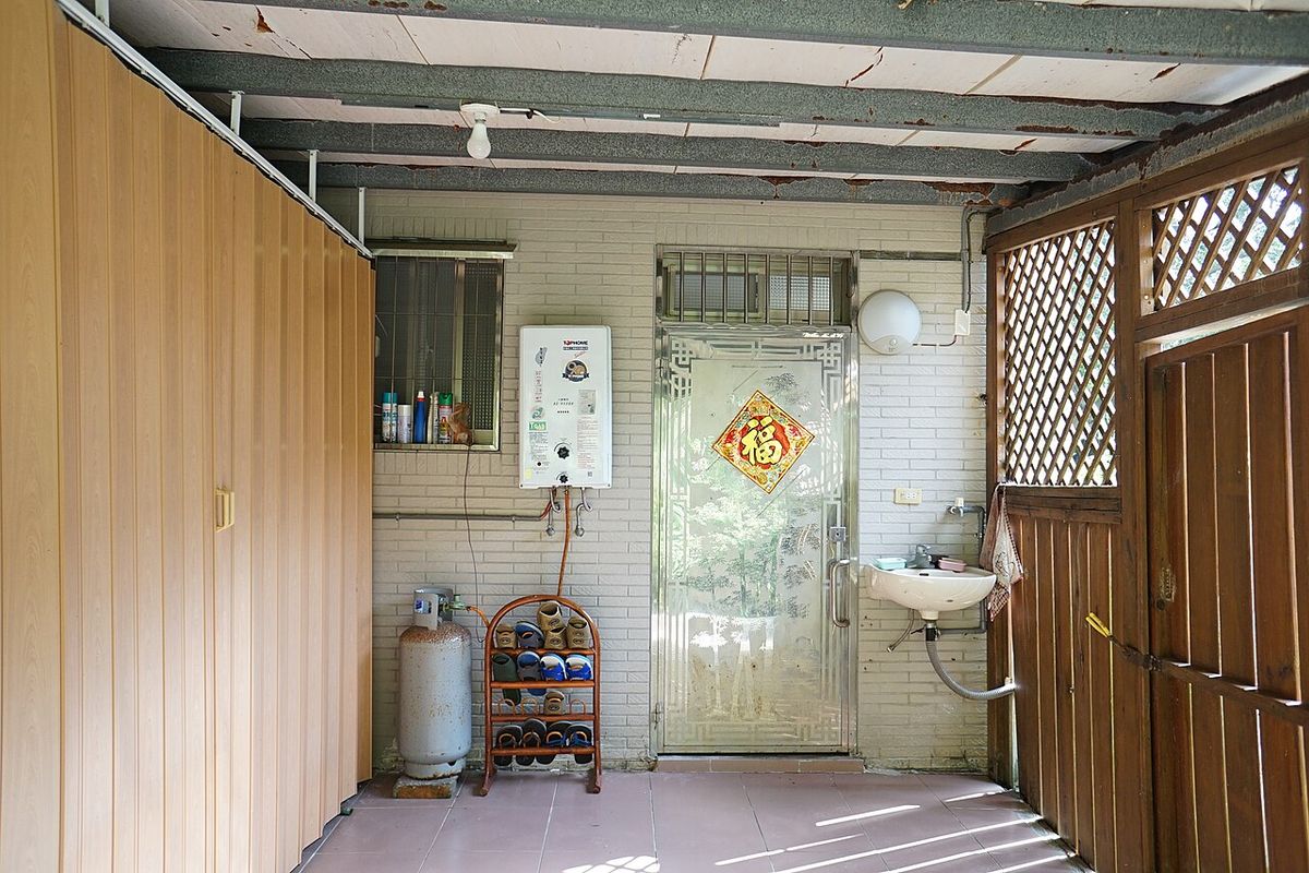 A white gas water heater mounted on a covered back porch in New Taipei, with a bottled gas cylinder on the floor below it