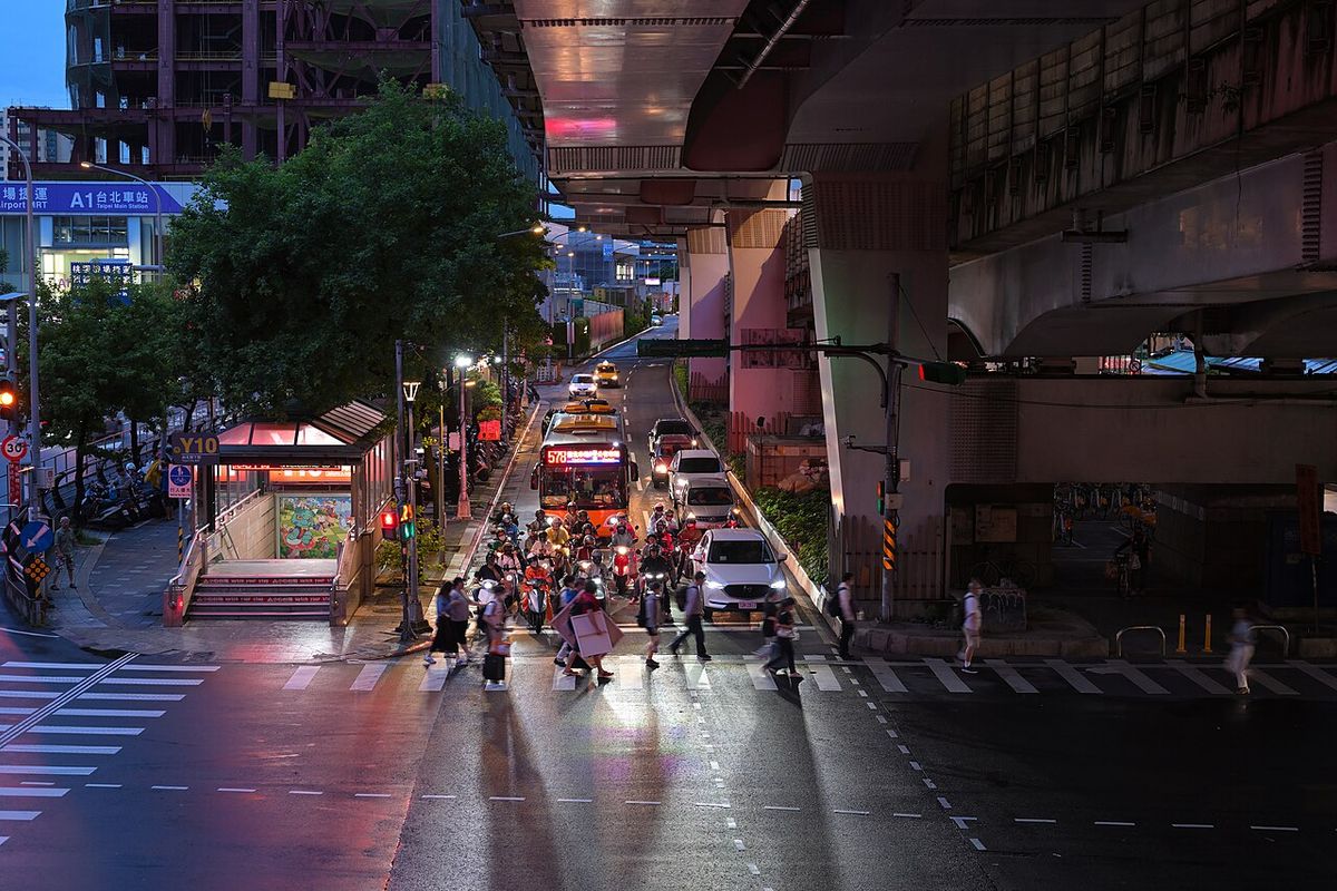 Pedestrians crossing at dusk under the elevated road near Taipei Main Station while scooters wait at the line