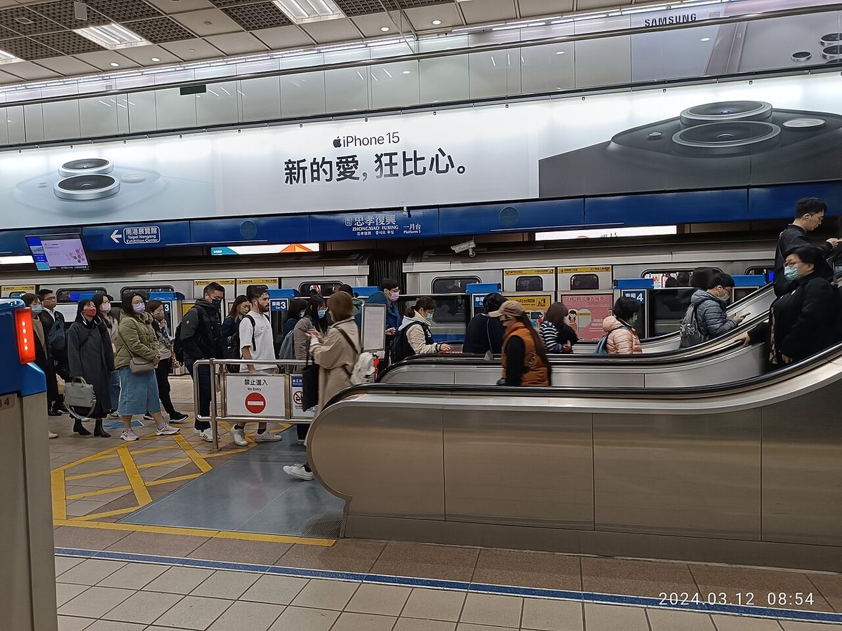 Commuters on a Taipei MRT platform at Zhongxiao Fuxing station, many looking at smartphones