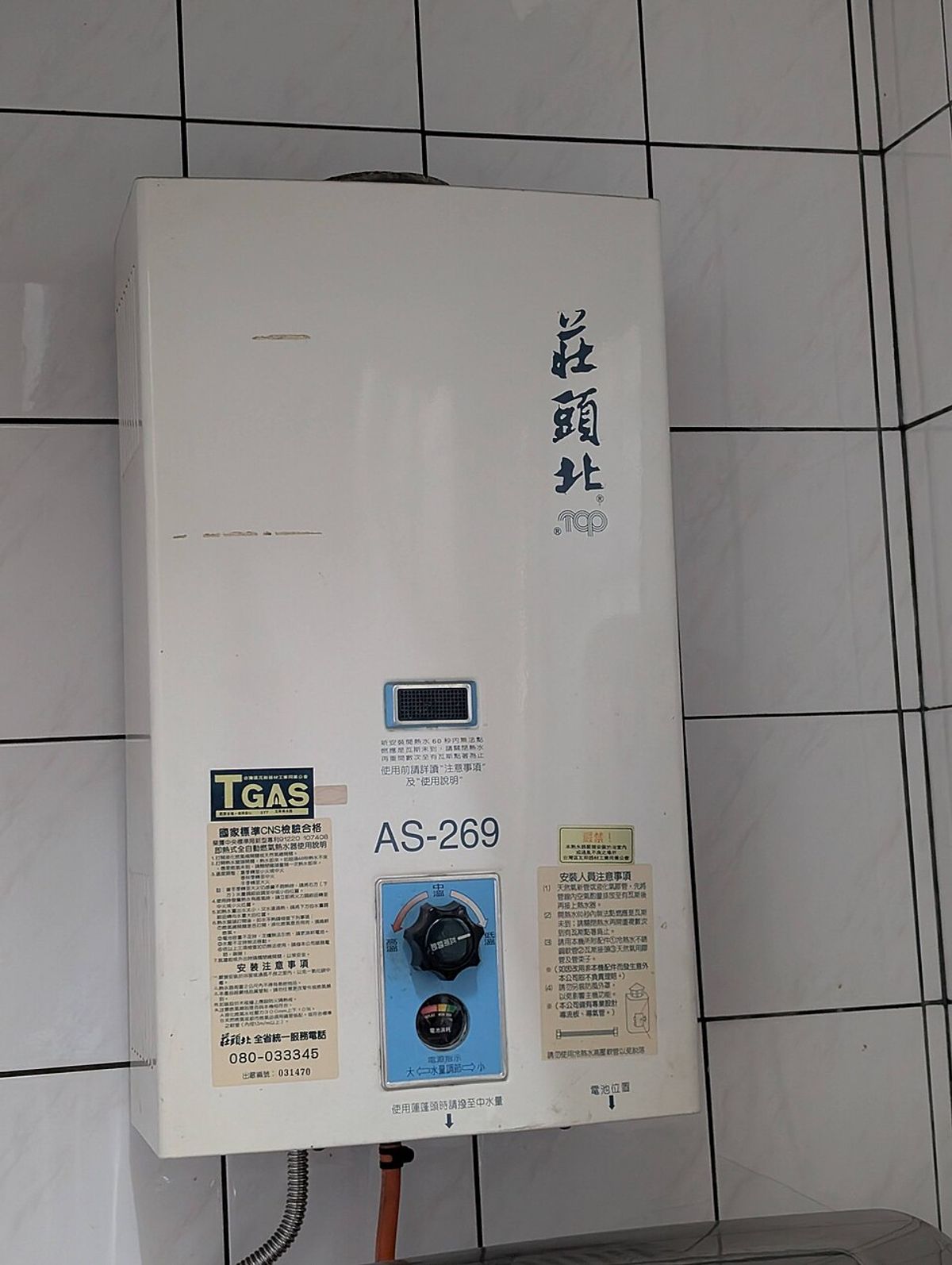 A gas water heater on a tiled wall showing the TGAS mark and CNS certification label on its front