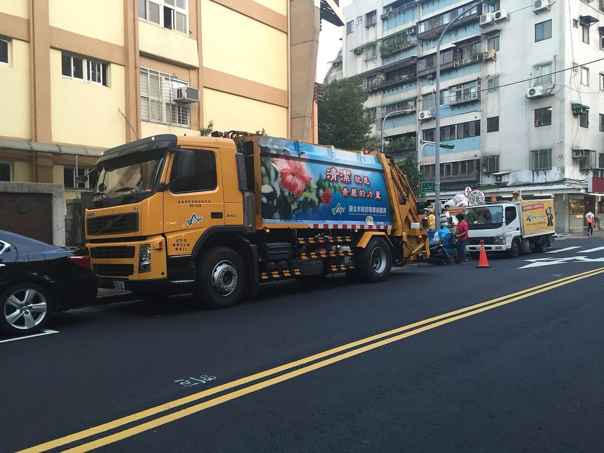 Two Taipei garbage trucks collecting trash on a narrow street lined with apartment buildings