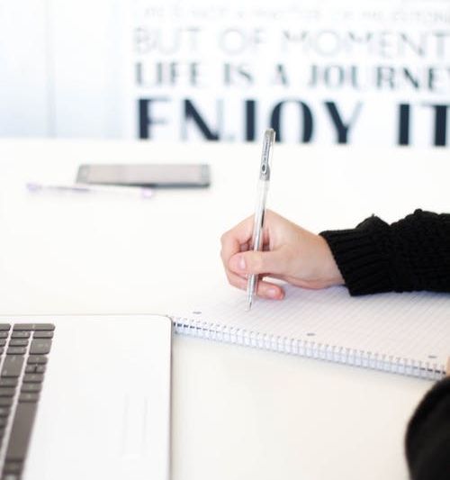 Image of a table with laptop and notebook, subject's hands are seen writing in the notebook