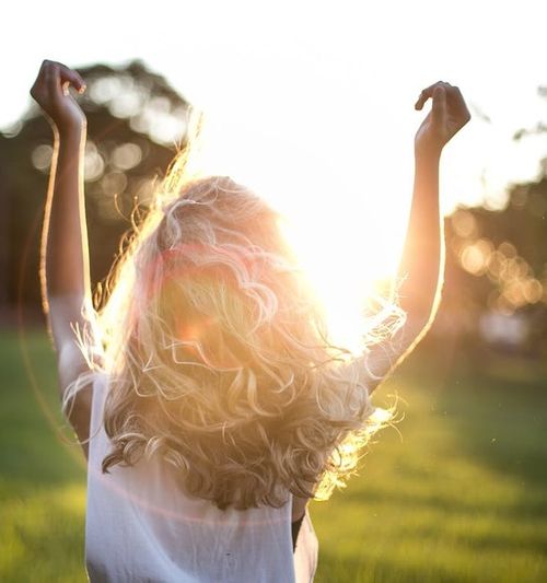 Image of a woman standing in the sunlight with back to camera and arms raised