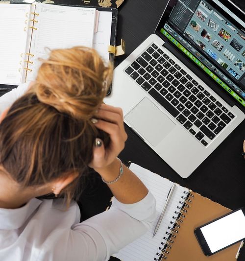 Image of a woman holding her head in her hands as she sits at her desk in front of a laptop and various notebooks and planners