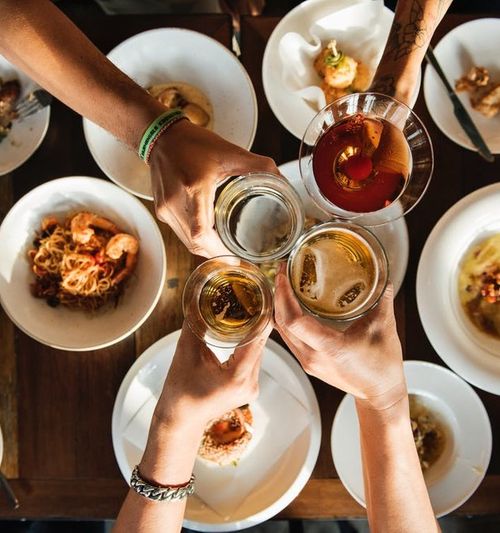 Image of four hands clicking their beers together, as seen from above over a table full of food