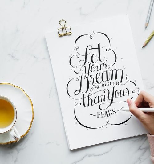 Image of a tabletop with a cup of tea and assorted items; a female hand is seen lettering a sign that reads "Let Your Dream Be Bigger Than Your Fears"