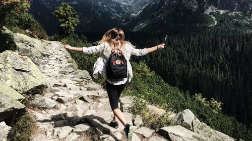 Image of a woman wearing a black backpack and casual clothing, seen from behind as she joyfully hikes through mountainous terrain 