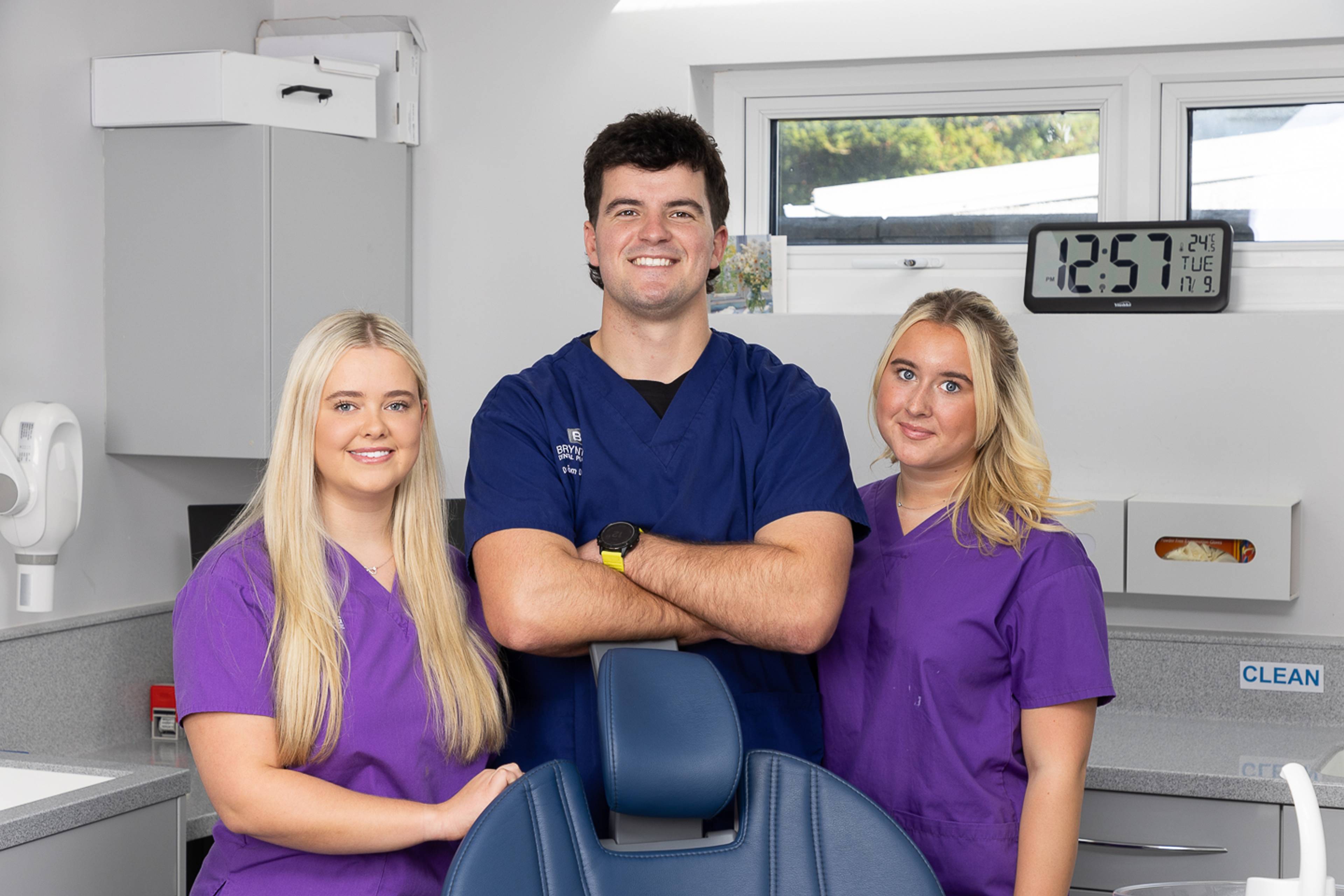 Roedd deintydd mewn prysgwydd glas a dwy nyrs mewn prysglwyni porffor yn sefyll y naill ochr a'r llall mewn deintyddfa ddeintyddol gwyn a llwyd plaen y tu ôl i gadair las