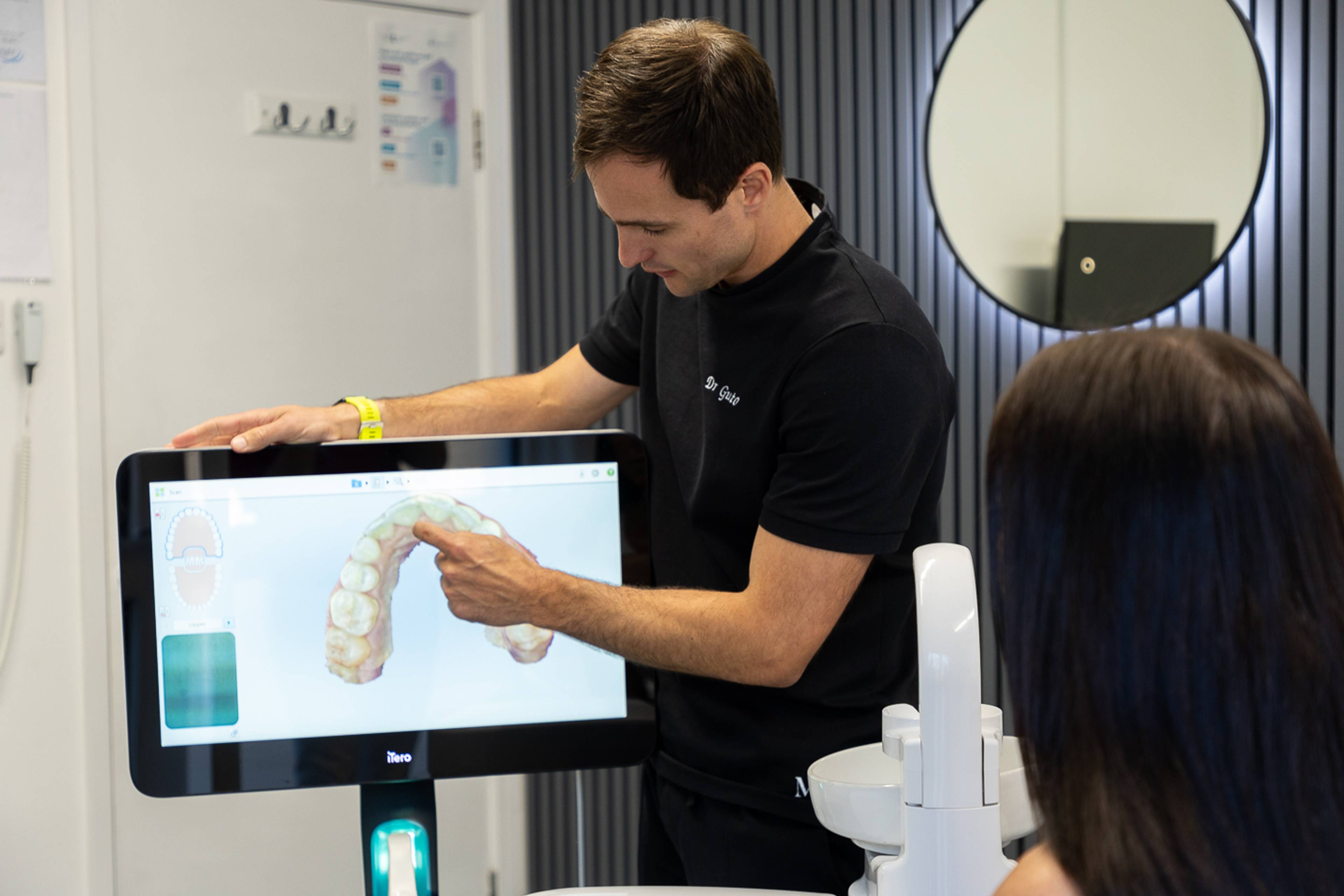 Dentist wearing black scrubs pointing to a screen showing a patient their 3D scan of their teeth