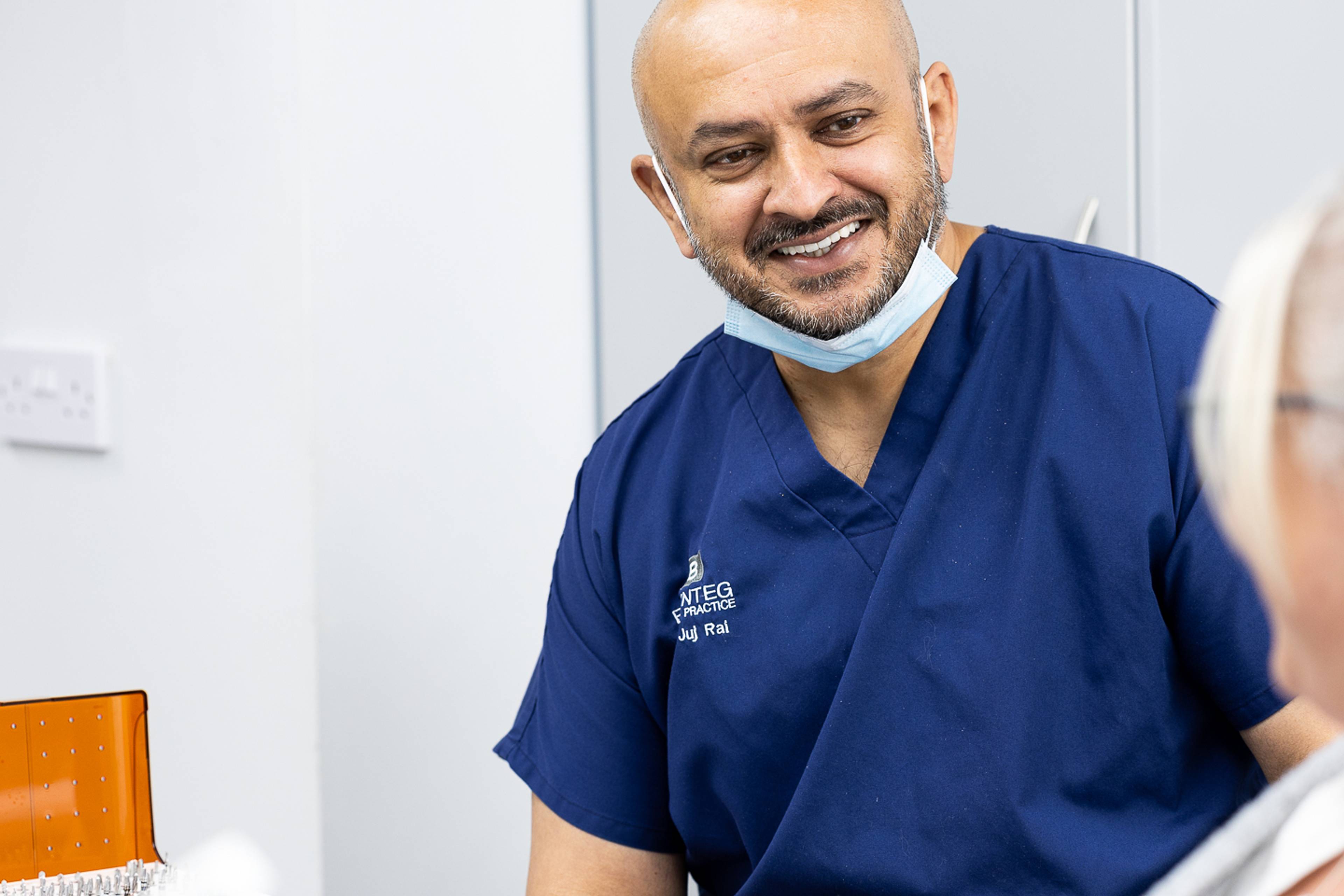 Deintydd mewn sgrwbiau glas gyda mwgwd o dan ên, yn gwenu tuag at y claf ar ymyl y llun yn y ddeintyddfa