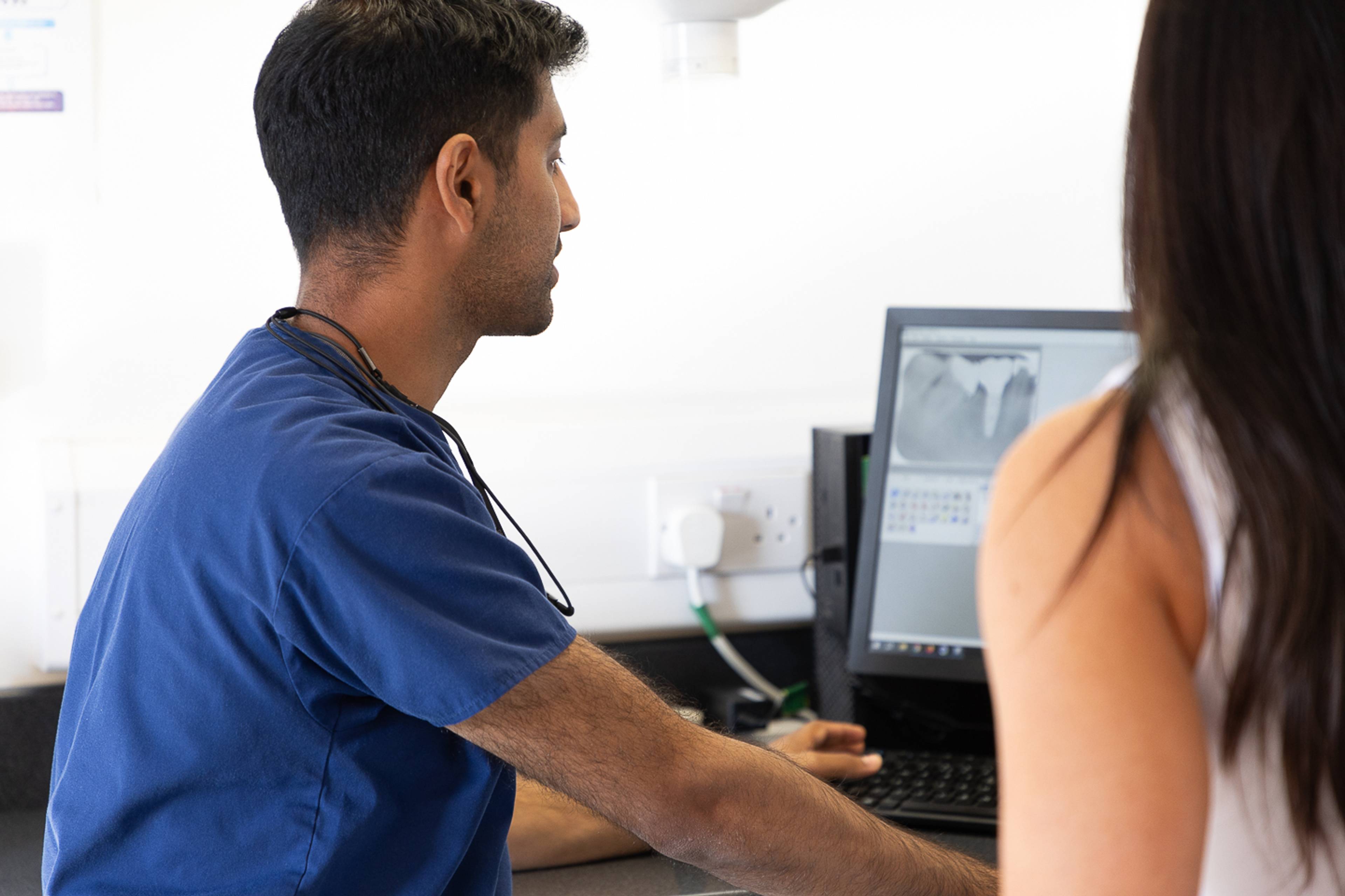 Deintydd mewn sgrwbiau glas yn wynebu i ffwrdd i sgrin y cyfrifiadur yn dangos eu pelydr-x i glaf