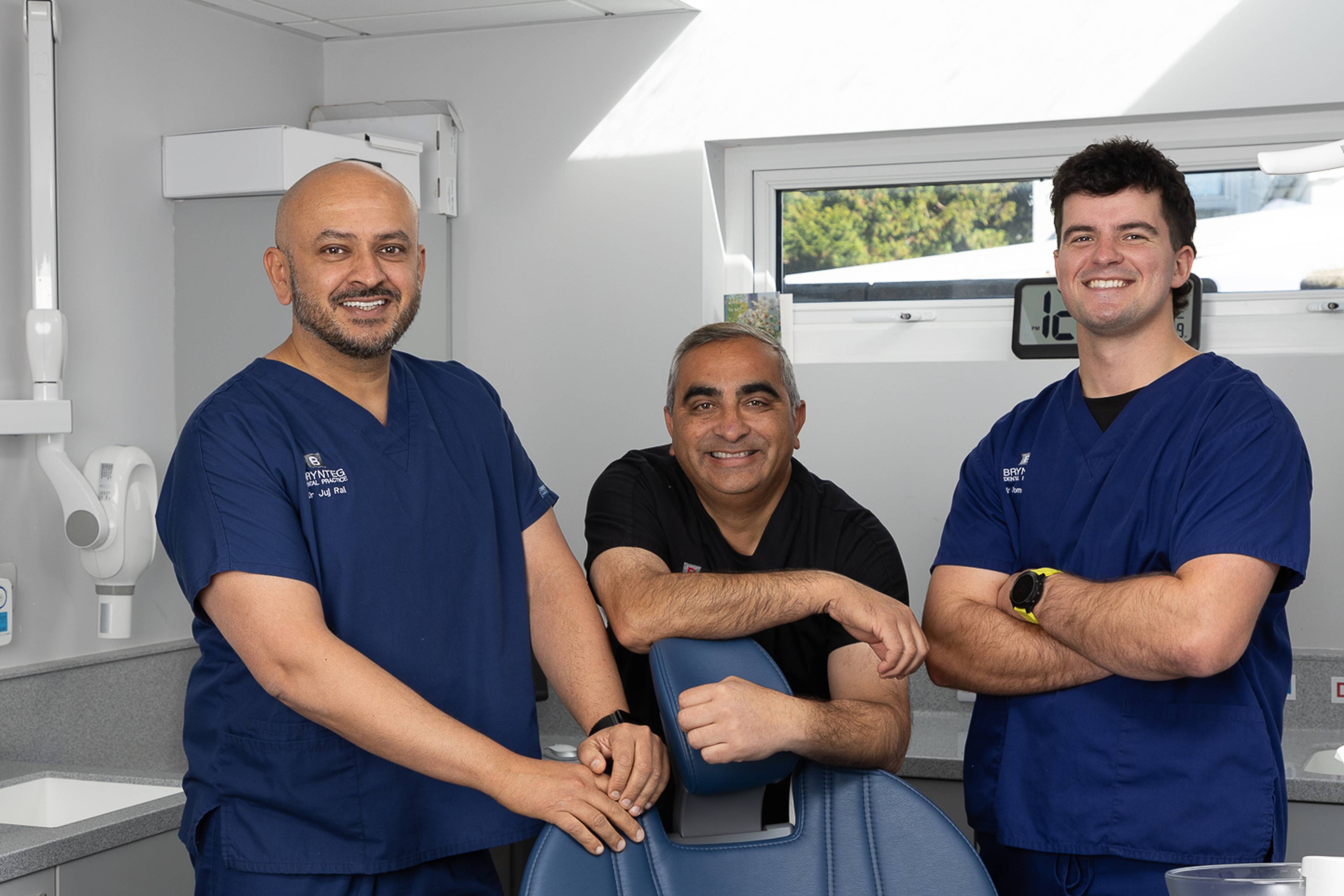 Dentists Juj, Nik & Tom standing around the dental chair, smiling towards camera
