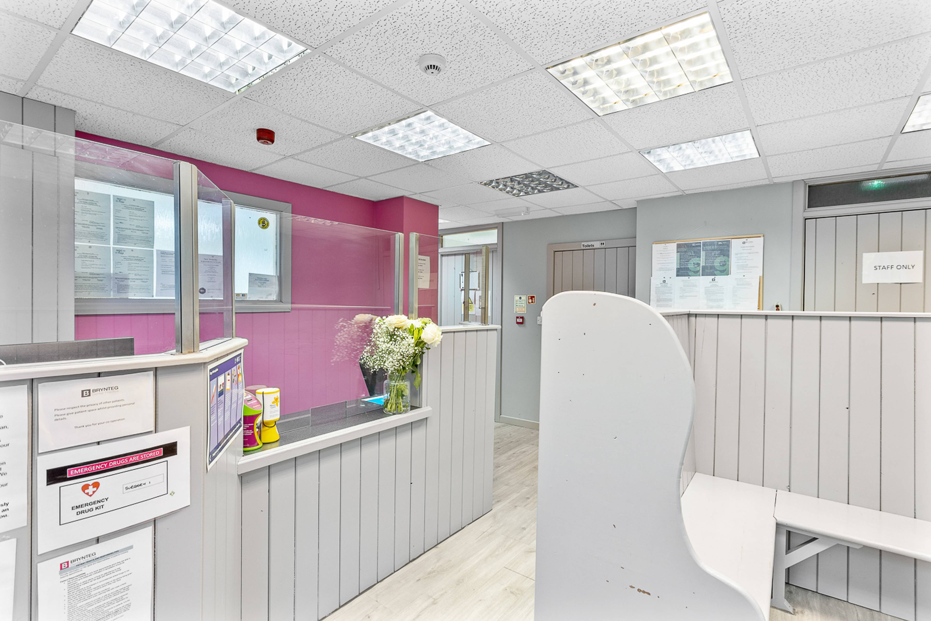 Deintyddol Brynteg Brynaman gyda seddau pren llwyd a wal binc y tu ôl i’r dderbynfa, sgrin a blodau wedi’u gosod wrth y ddesg flaen