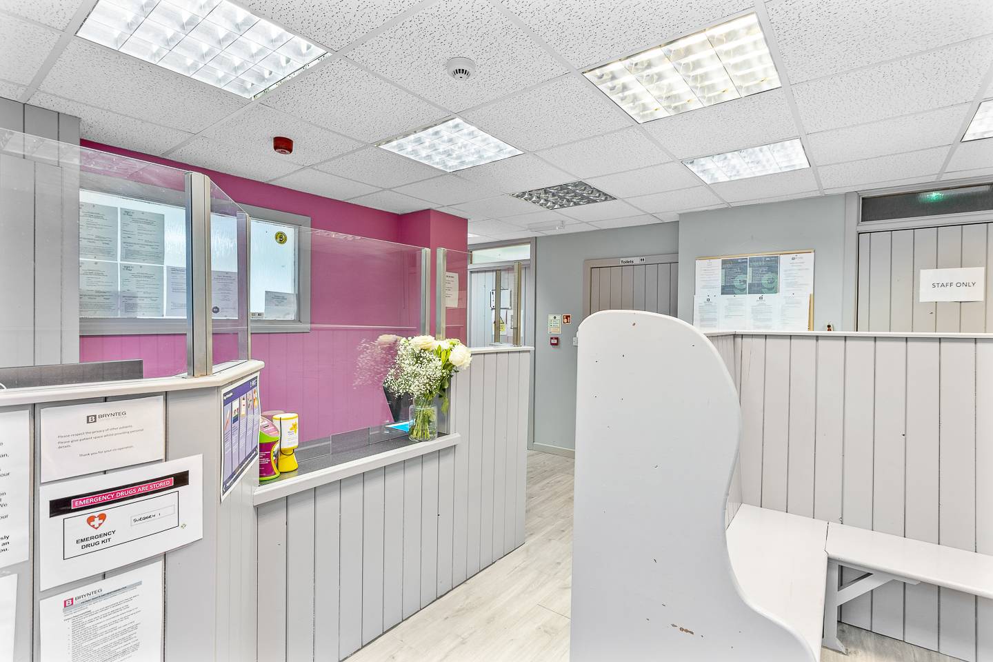 Deintyddol Brynteg Brynaman gyda seddau pren llwyd a wal binc y tu ôl i’r dderbynfa, sgrin a blodau wedi’u gosod wrth y ddesg flaen