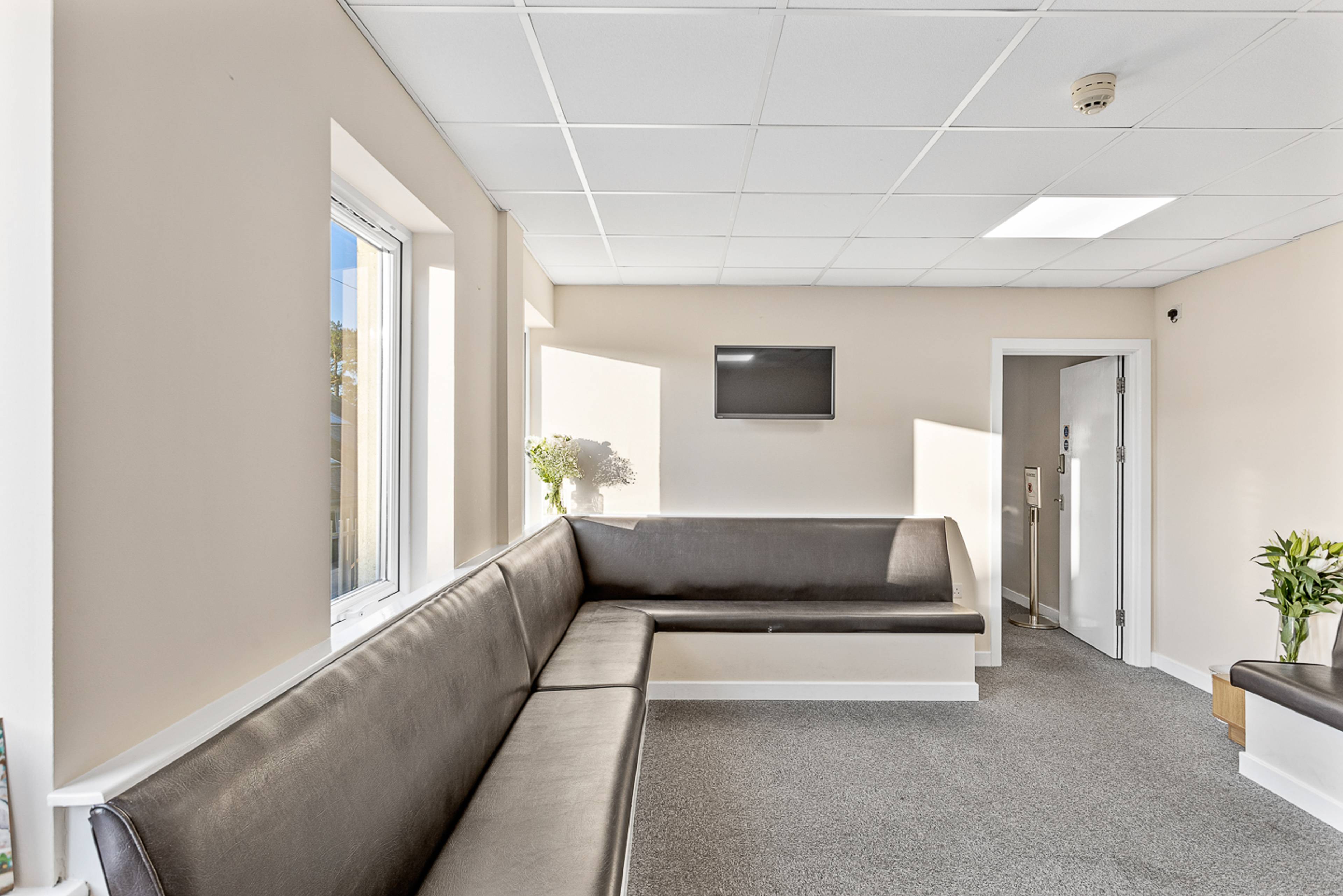 Ystafell aros Deintyddol Brynteg Caerfyrddin gyda waliau hufen, seddi soffa llwyd ar hyd waliau, golau yn dod i mewn o ffenestr fawr a sgrin deledu ar y wal