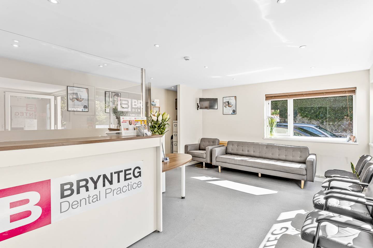 Ystafell aros Deintyddol Brynteg Dinbych-y-pysgod gyda soffas llwyd a waliau hufen, ffenestri mawr, golau a modern