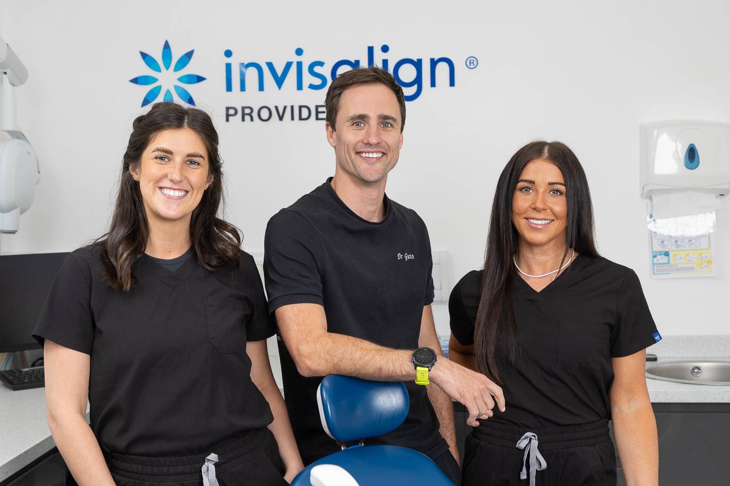 Dental team wearing black scrubs in front of Invisalign wall sign in dental surgery