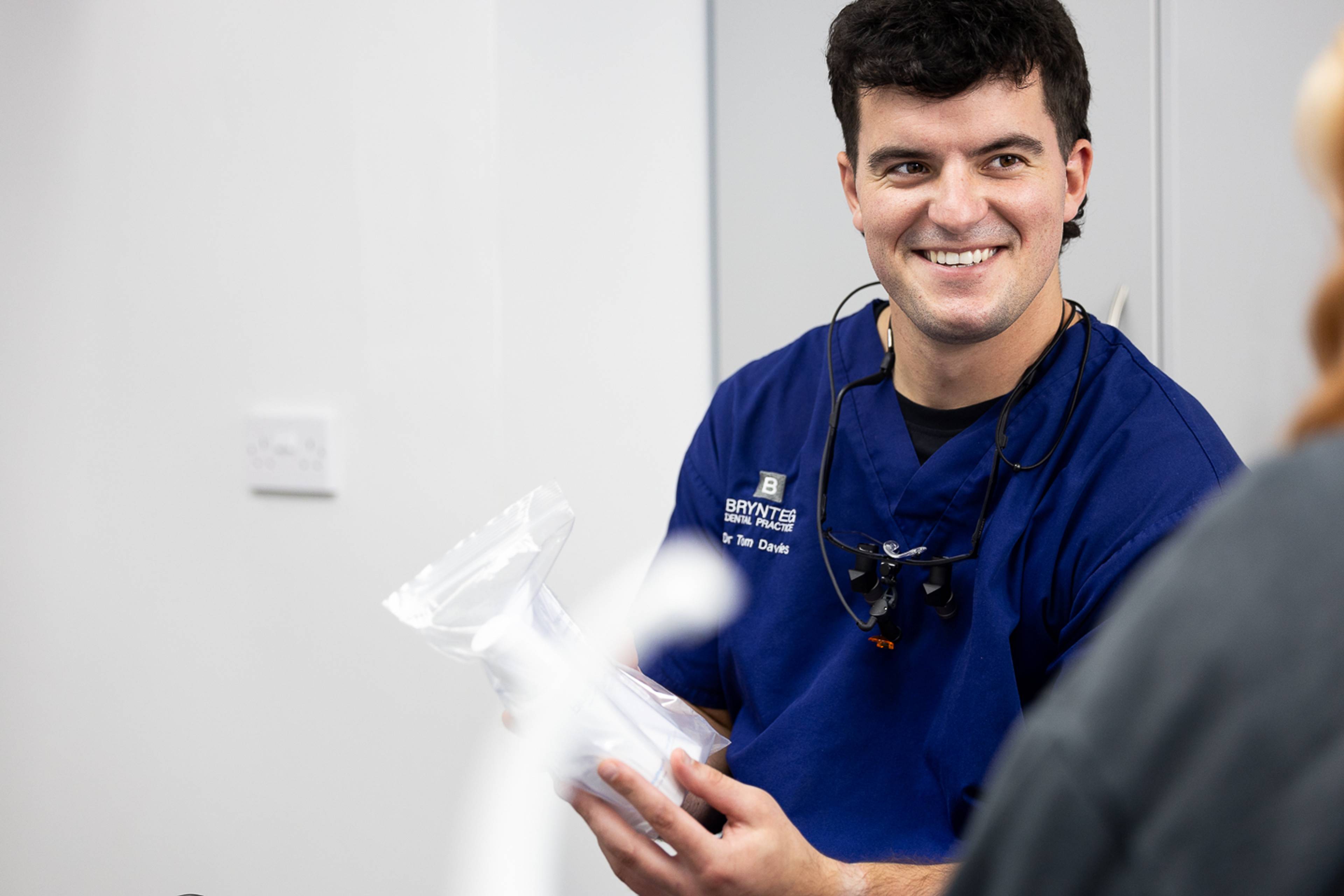 Male dentist in dark blue scrubs holding a dental model whilst smiling at the patient. 