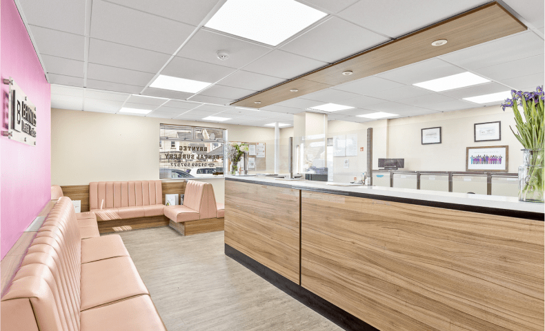 Ystafell aros Deintyddol Brynteg Rhydaman, gyda soffas cefn uchel llwydfelyn, wal â brand pinc a derbynfa bren modern yng nghanol yr ystafell