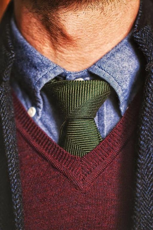 Una fotografía close-up del atuendo de un hombre que complementa una corbata verde oliva con un suéter rojo granate.