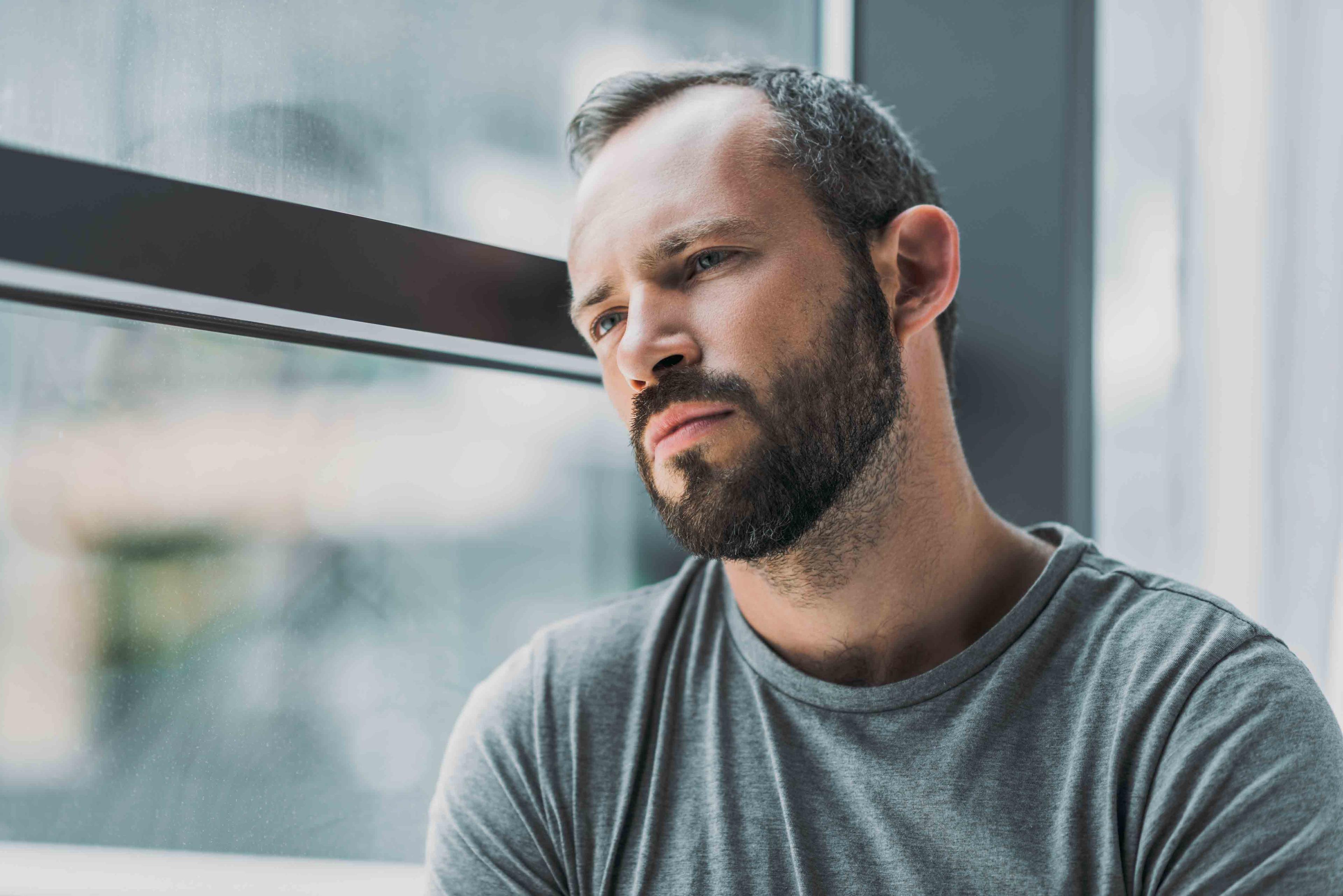 Sad bearded brunette man leaning against a window and looking away from the camera.