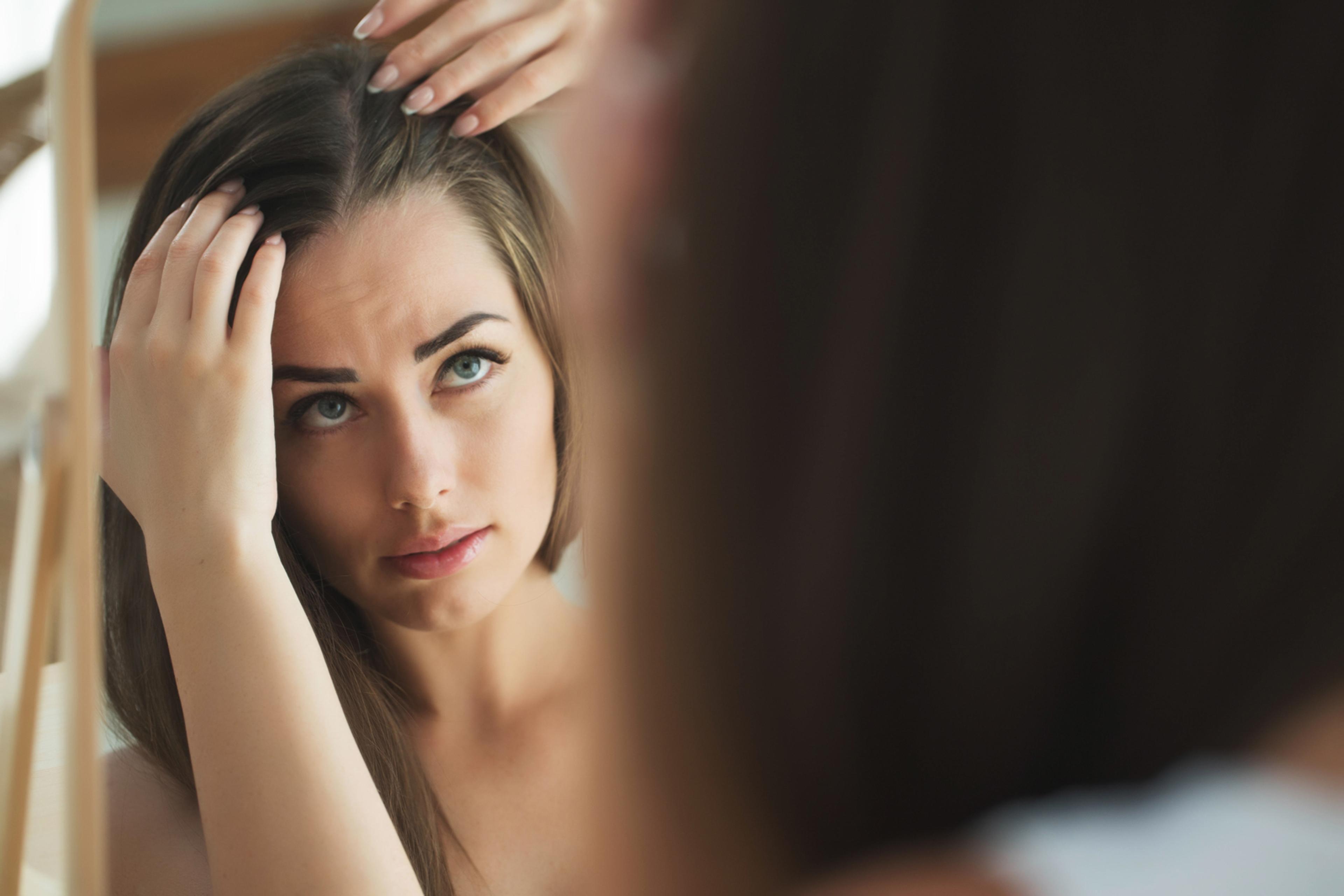 Concerned young brunette woman looking into mirror and examining her hairline for shines of thinning.