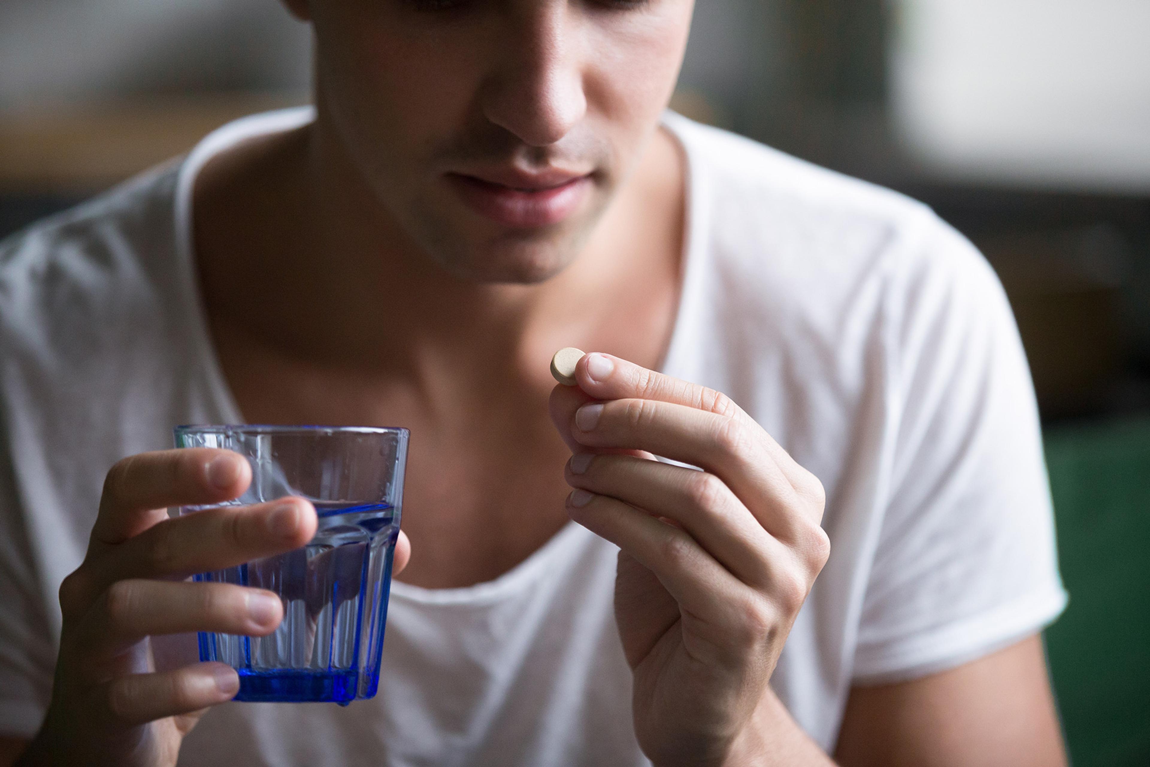 Young man holding glass of water in one hand and pill in the other, and examining the pill intently.